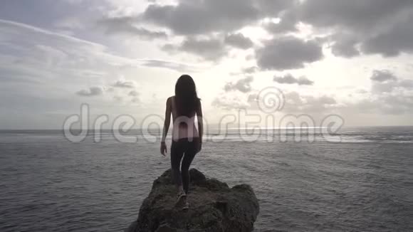 站在岩石上望着大海举手的年轻女子视频的预览图