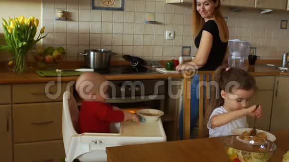 可爱的微笑家庭主妇正在做饭同时在厨房里喂两个孩子女人窥视视频的预览图