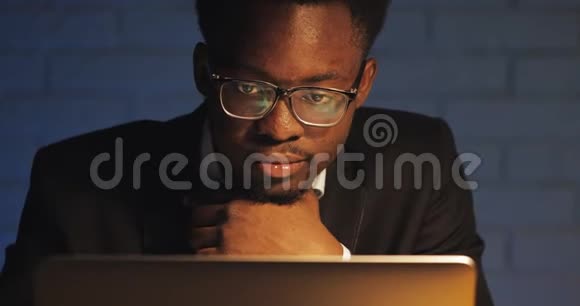 疲惫的年轻黑人商人在夜间办公室用笔记本电脑工作在黑暗房间加班的自由职业者视频的预览图