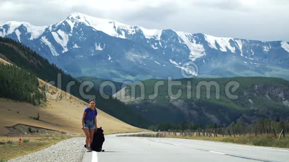 年轻的旅游妇女搭便车正在山路上拉一个沉重的背包那里有雪山视频的预览图