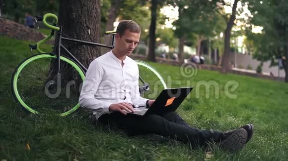 穿白衬衫的年轻成功商人一名男子坐在草地上在绿茵上的城市公园里用笔记本电脑工作视频的预览图