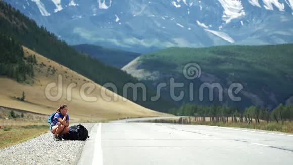 一个搭便车的女孩坐在背包旁在山路上等车那里有雪山视频的预览图