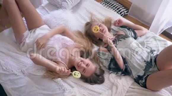 舒适的气氛穿着丝绸睡衣的姐妹们吃着甜棒棒糖躺在家里的床上聊天视频的预览图