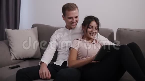 年轻人坐着女人随意地躺在家里的灰色沙发上看着平板电脑一起度过时光视频的预览图