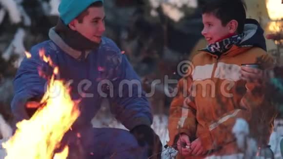 冬季森林年轻的一家人坐在火边的树林里喝着泰莫斯的热饮和炸香肠视频的预览图