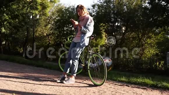 年轻漂亮的女孩靠在城市公园里的自行车上看着她的手机和Smolong穿着明亮的粉红色和蓝色视频的预览图