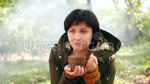 一位女游客在炉火旁的秋天花园里从杯子里喝热茶或咖啡漂亮的女孩在发抖视频的预览图