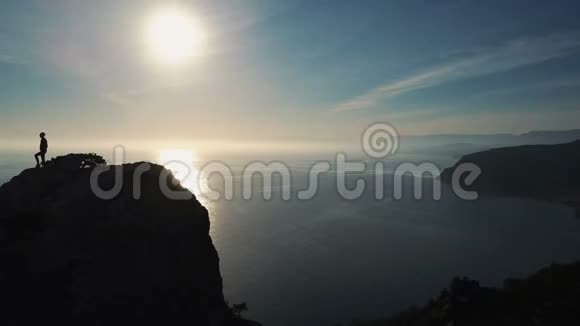 夕阳西下一位年轻女子站在山顶上靠海的剪影女士视频的预览图
