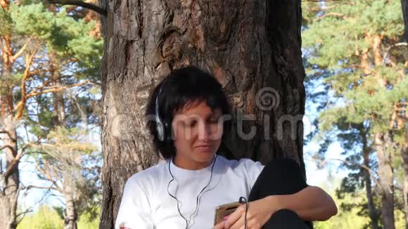 一个戴耳机的女孩的特写一位女游客坐在树旁在智能手机上听音乐摇着头视频的预览图