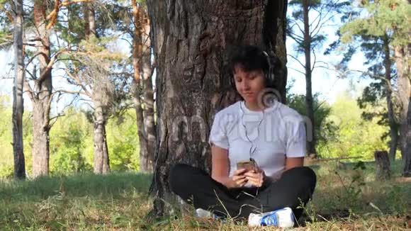 一个女孩坐在带智能手机的耳机里在公园的背景下听音乐互联网音乐和视频的预览图