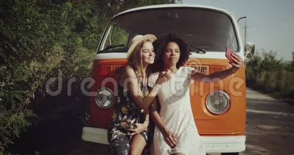 自拍时光在复古橙色巴士旁边两个女孩金发和非洲卷发穿着一个视频的预览图