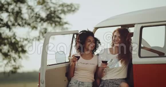 两个年轻的女人野餐的时候喝着酒站在复古的公共汽车旁边卷发心情很好视频的预览图
