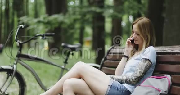 一个有纹身的年轻女人坐在公园的长凳上在手机上说话视频的预览图