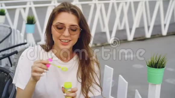 戴着太阳镜的漂亮年轻女人正坐在户外笑着吹肥皂泡夏日休闲青春视频的预览图