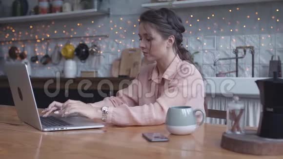 集中精力的年轻女子坐在厨房的木桌旁在她的银色笔记本电脑上用茶或视频的预览图