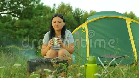 一个女人坐在帐篷附近喝茶在烤架上煮肉视频的预览图