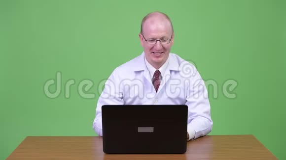 快乐成熟的秃头医生使用笔记本电脑在木制桌子上得到好消息视频的预览图