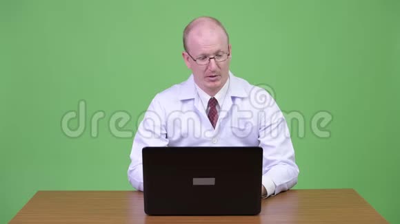 一位衣着成熟的秃头医生对着笔记本电脑和木桌争吵视频的预览图