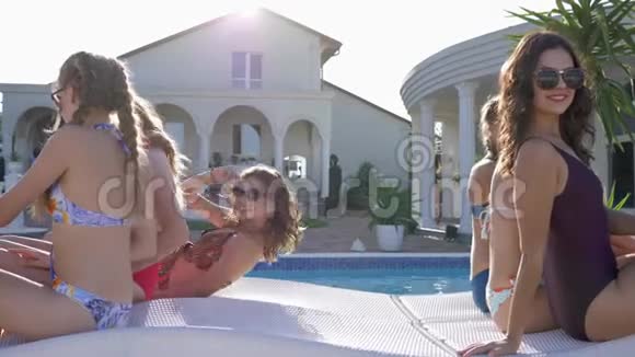 泳池派对穿着泳衣和太阳镜的快乐女朋友同时躺在游泳池附近的躺椅上视频的预览图