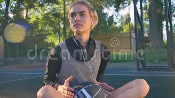 漂亮的年轻女子坐在篮球场上通过耳机听音乐拿着球打开空气镜头视频的预览图