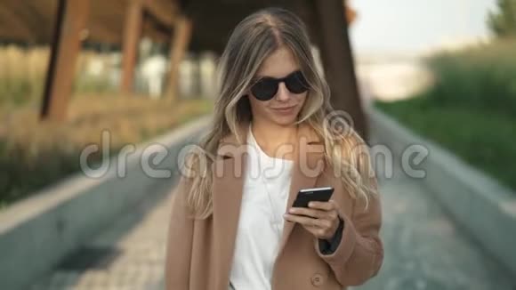 在秋天公园散步的金发女郎微笑着看着智能手机和打字视频的预览图
