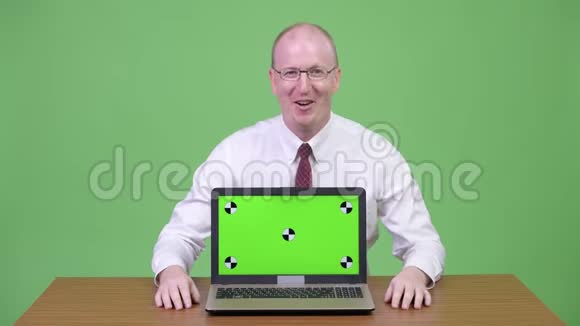 快乐成熟的秃头商人在木制桌子上展示笔记本电脑视频的预览图