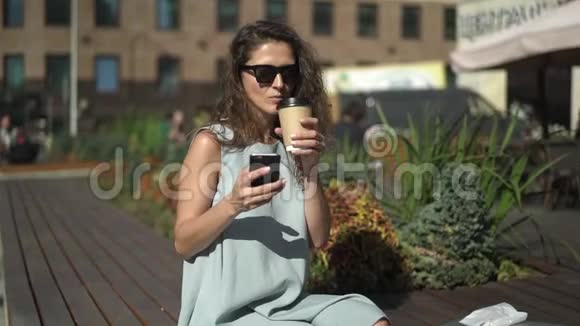 美丽的年轻女子喝着咖啡在户外咖啡馆附近看着电话视频的预览图