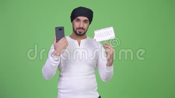 年轻的留着胡子的印度男人拿着手机和自拍纸标志视频的预览图