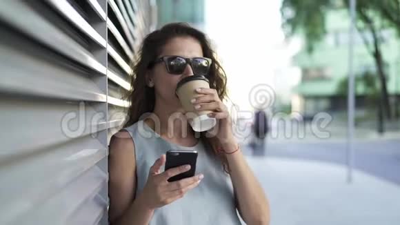 在夏季城市的灰色墙壁附近喝咖啡和上网的女人视频的预览图