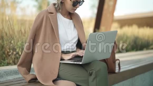 穿着米色外套的金发女人在秋天公园用笔记本电脑工作视频的预览图