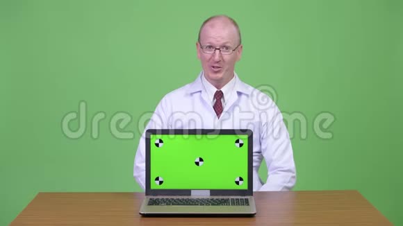 快乐成熟的秃头医生一边说话一边拿着笔记本电脑对着木桌视频的预览图