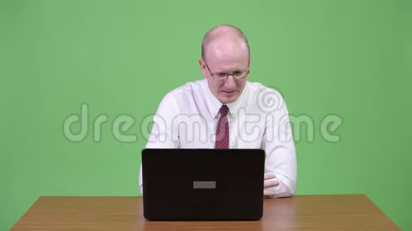 穿着成熟的秃头商人用手提电脑在木制桌子上听到坏消息视频的预览图
