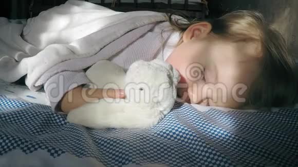 美丽的小女孩金发睡在床上被阳光照亮带着一只泰迪兔子视频的预览图