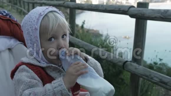 穿着连帽衫和红色针织背心的小女孩喝牛奶或塑料瓶中的乳制品坐在婴儿车上视频的预览图