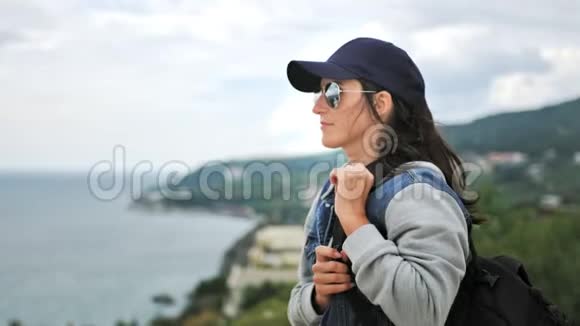 时尚的徒步旅行者戴着墨镜和帽子站在山顶欣赏美丽的海景视频的预览图