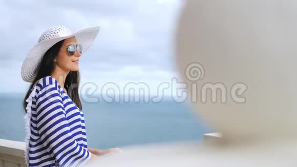 在阳台或露台上欣赏大海和天空的优雅女人戴着帽子和太阳镜视频的预览图