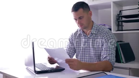在办公室工作时一个悲伤的年轻人疲倦地看着文件然后在笔记本电脑屏幕上视频的预览图