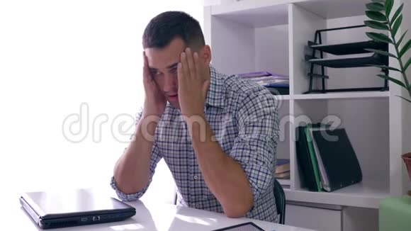 一位头痛的商人在办公室工作时坐在桌子上手里拿着一台关闭的笔记本电脑视频的预览图