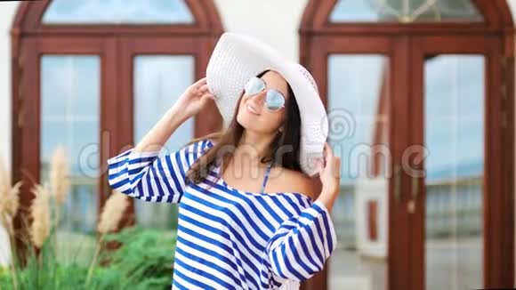 美丽的女游客戴着帽子太阳镜和条纹连衣裙玩得很开心摆姿势视频的预览图