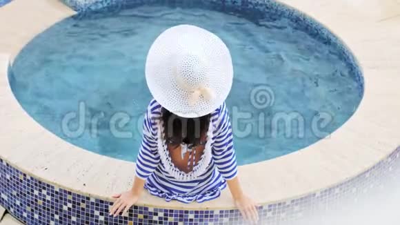 高角度后景戴帽子的优雅女人带蝴蝶结坐在游泳池或按摩浴缸边缘视频的预览图