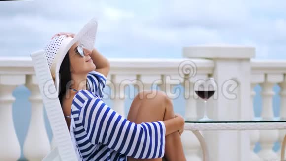 优雅的女士戴着太阳镜戴着帽子坐在阳台上交叉双手抱着腿坐在椅子上视频的预览图