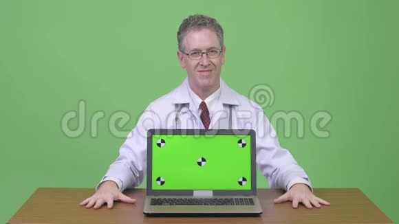 快乐的成熟男人医生展示笔记本电脑用大拇指对着木桌视频的预览图