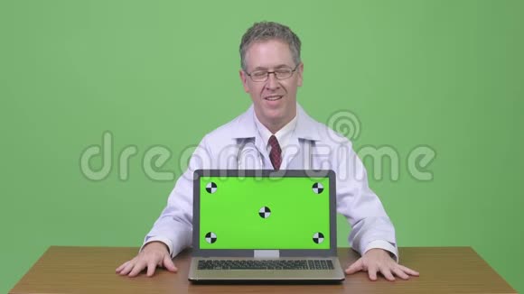 快乐的成熟男人医生在木制桌子上展示笔记本电脑视频的预览图