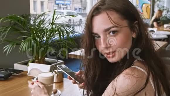 快乐女孩用手机应用程序在咖啡馆里微笑地拍摄咖啡手机上美丽的年轻休闲女性视频的预览图