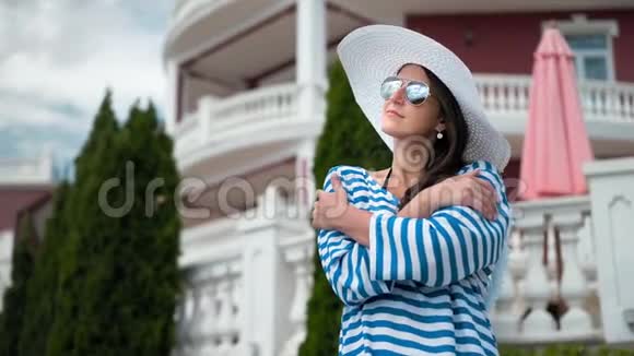 戴墨镜和戴帽子的低角度优雅女人双手交叉站立微笑着享受视频的预览图