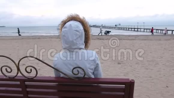 穿着白色羽绒服的女人坐在海滩上的长凳上看着远处海冬后景视频的预览图