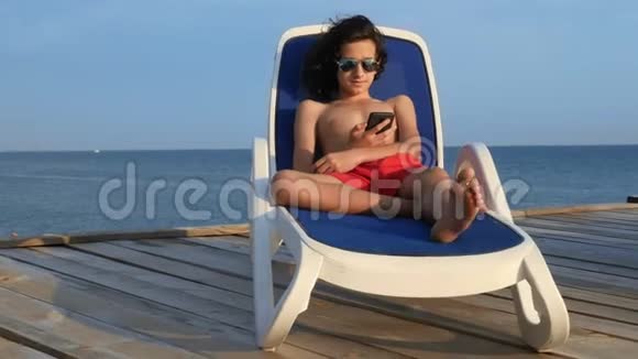 美丽的少年男孩卷曲的黑发躺在甲板上的椅子上在一个木制的海面上使用智能手机视频的预览图