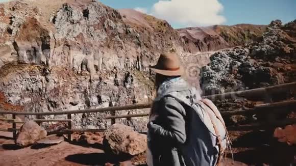 年轻的女性旅行博主带着背包戴着帽子从维苏威火山徒步下来拍摄阳光明媚的火山口视频的预览图