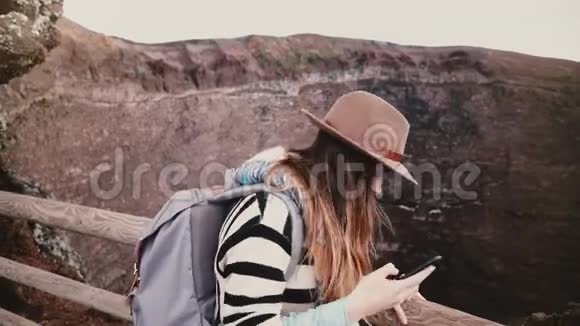 年轻漂亮的学生女孩带着背包微笑在维苏威火山火山口边缘拍摄智能手机自拍照片视频的预览图