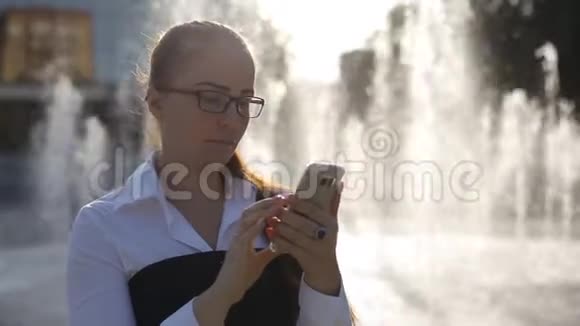 白人年轻女子戴着光学眼镜出现在公园喷泉附近手里拿着智能手机视频的预览图